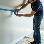 A person carefully applying blue painter's tape along a wall edge before painting a room