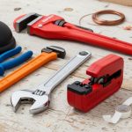 A organized set of essential home plumbing tools including wrenches, a plunger, pipe cutter, and PTFE tape laid out on a workbench