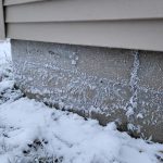 Snow-covered yard with an exposed foundation wall showing frost and ice