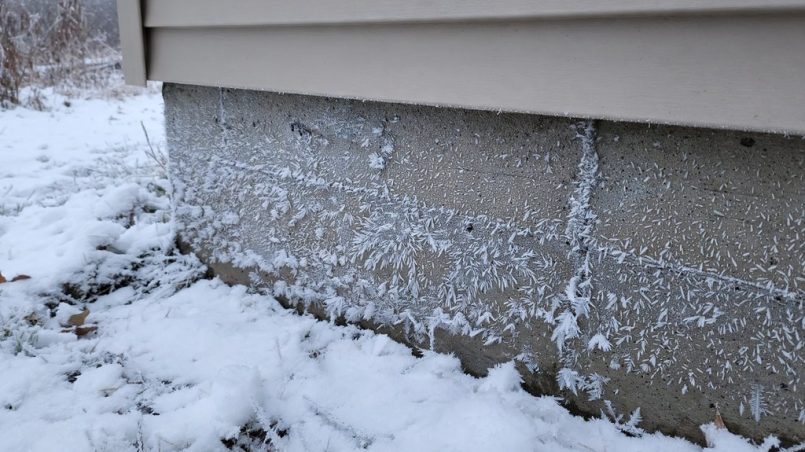 Snow-covered yard with an exposed foundation wall showing frost and ice